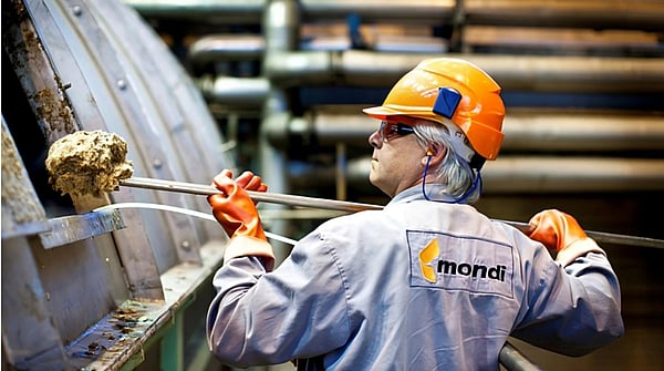 A Mondi employee wearing grey overalls, safety goggles and an orange hardhat picks up residues from wood fibres, chemicals and other substances from a large machine.